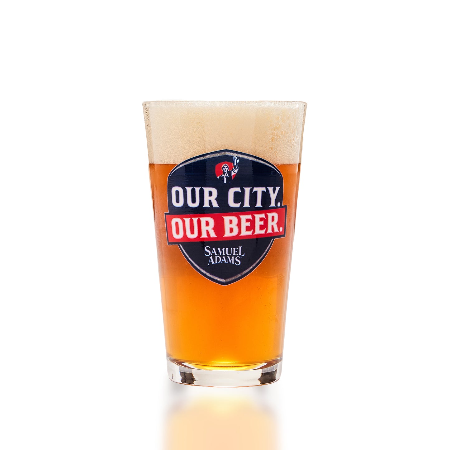 A clear pint glass filled with amber beer and topped with white foam, featuring a prominent Samuel Adams 'Our City Our Beer' logo on the front, set against a clean white background.