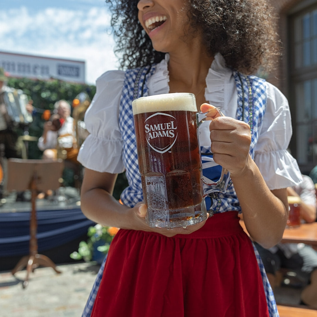 Woman in traditional German outfit holding a Samuel Adams beer mug outdoors.