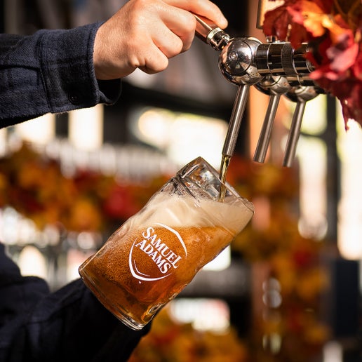 A half liter Octoberfest Glass Stein with a white Sam Adams logo on it, modeled after authentic Bavarian beer glassware being held by a person dispensing a beer on tap. Glass is made of dimpled heavy duty glass.