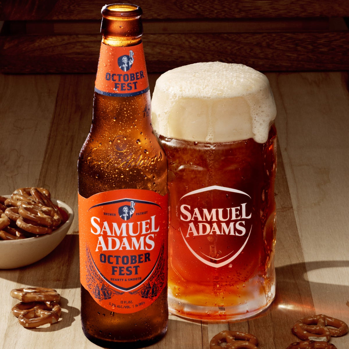 A bottle and a frothy glass of Samuel Adams Octoberfest beer on a wooden table, with pretzels scattered around and a bowl of pretzels in the background.