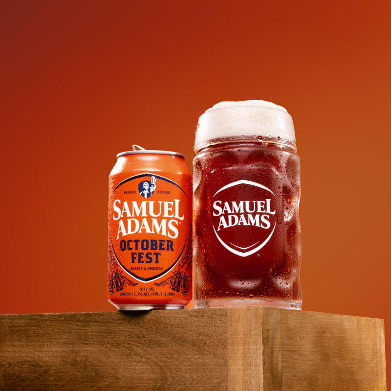 A Samuel Adams Octoberfest orange beverage can and a matching frothy glass mug with the Samuel Adams logo, displayed on a wooden block against a warm orange gradient background.