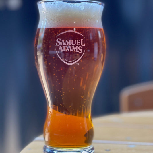 A 22-ounce pint glass filled with beer with the classic Samuel Adams shield logo printed on the front in white.