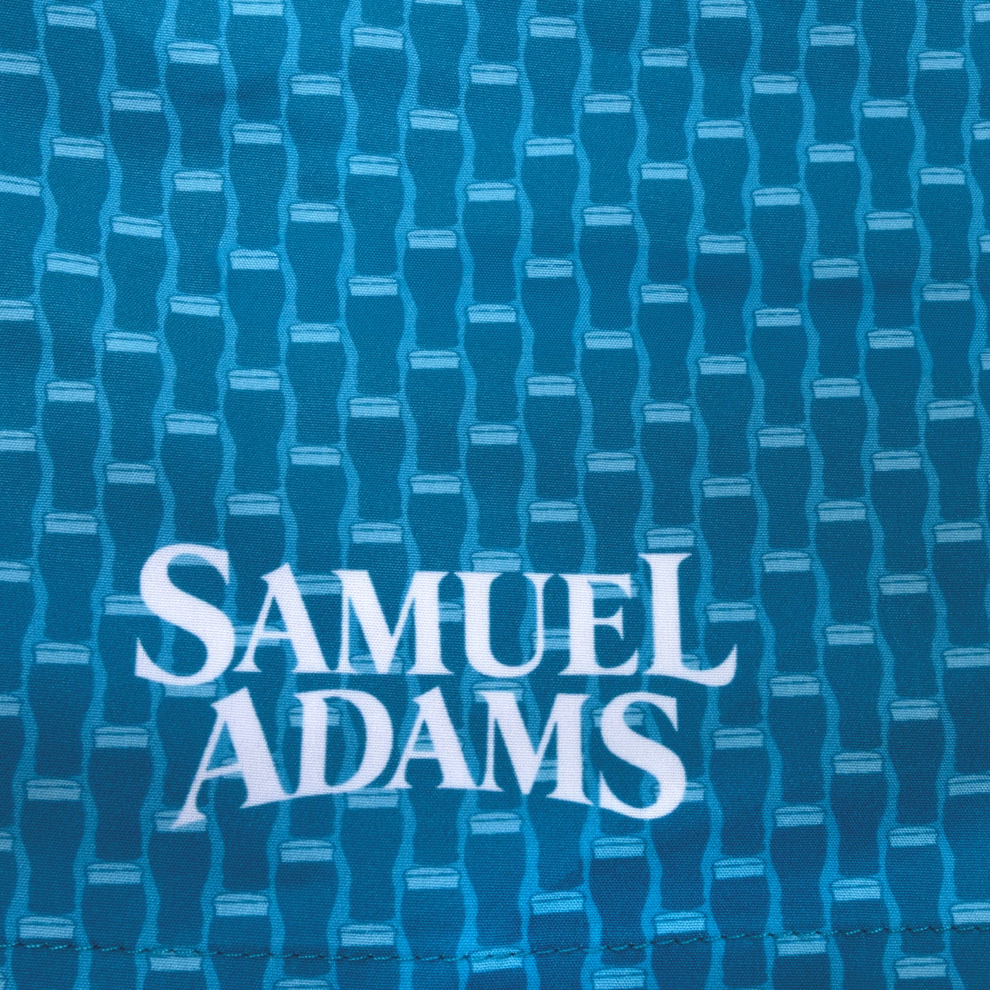 Close-up of Samuel Adams swim trunks featuring blue pint glass pattern fabric and white logo print on quick-dry material