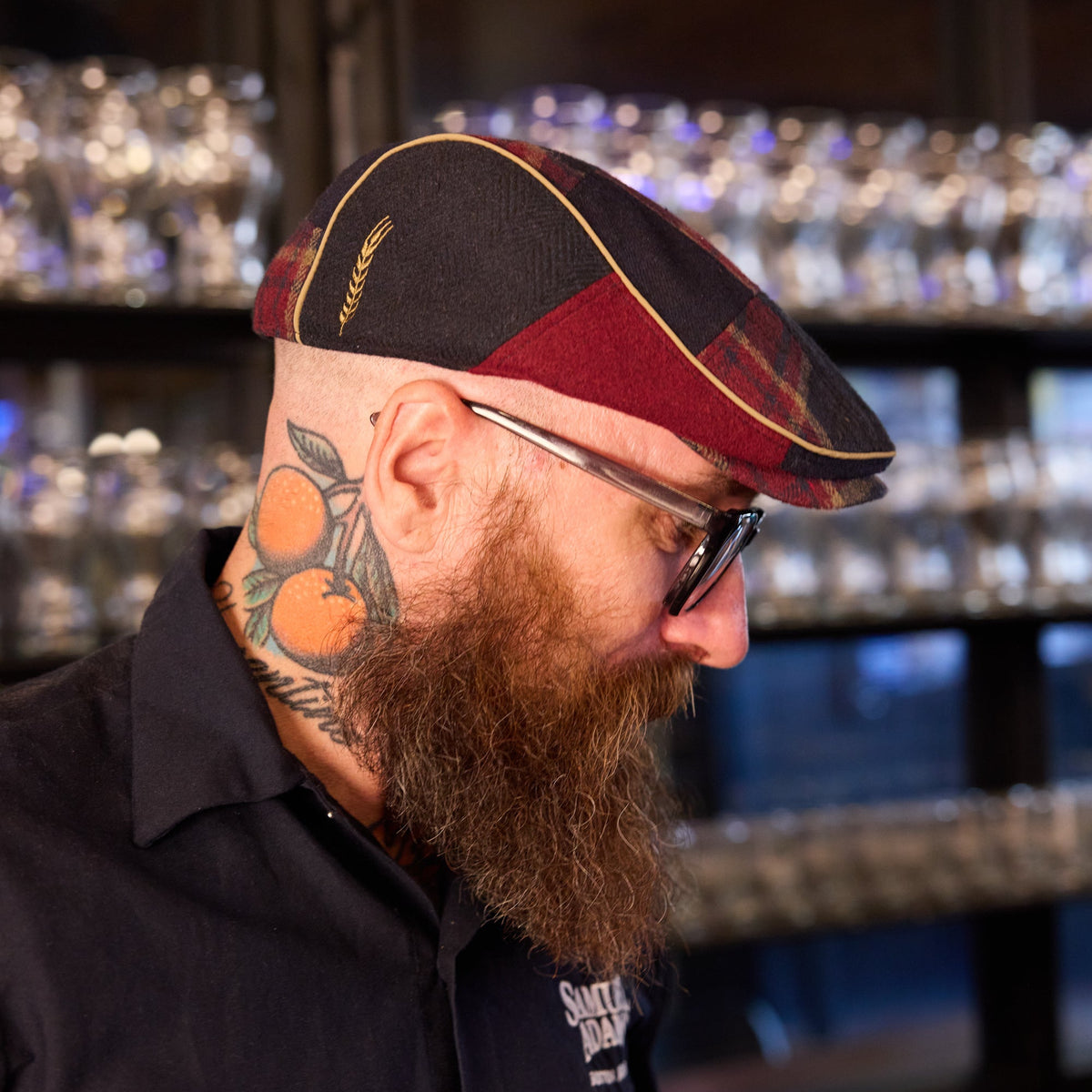 A man with a full beard and tattoos wearing a black and maroon plaid flat cap with gold piping, glasses, and a dark shirt, photographed in a dimly lit bar setting.