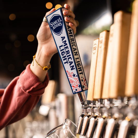 A Samuel Adams American Light tap handle with a white background, blue trim/details, and red details being poured