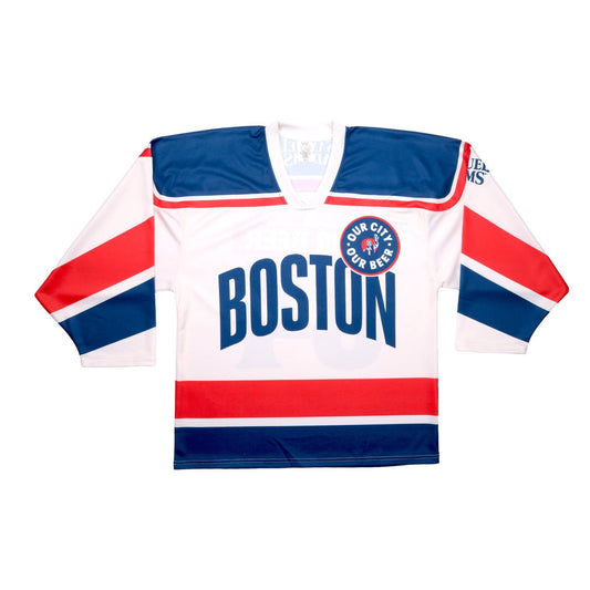 Base of jersey is white with blue and red patterns on the arms, waist, and shoulders. Centered "Boston" is written in blue, with a circle badge which features "Our City, Our Beer" and a red shield with Sam Adams hoisting a beer in the center of it. The Samuel Adams logo is featured in blue on the upper left arm.