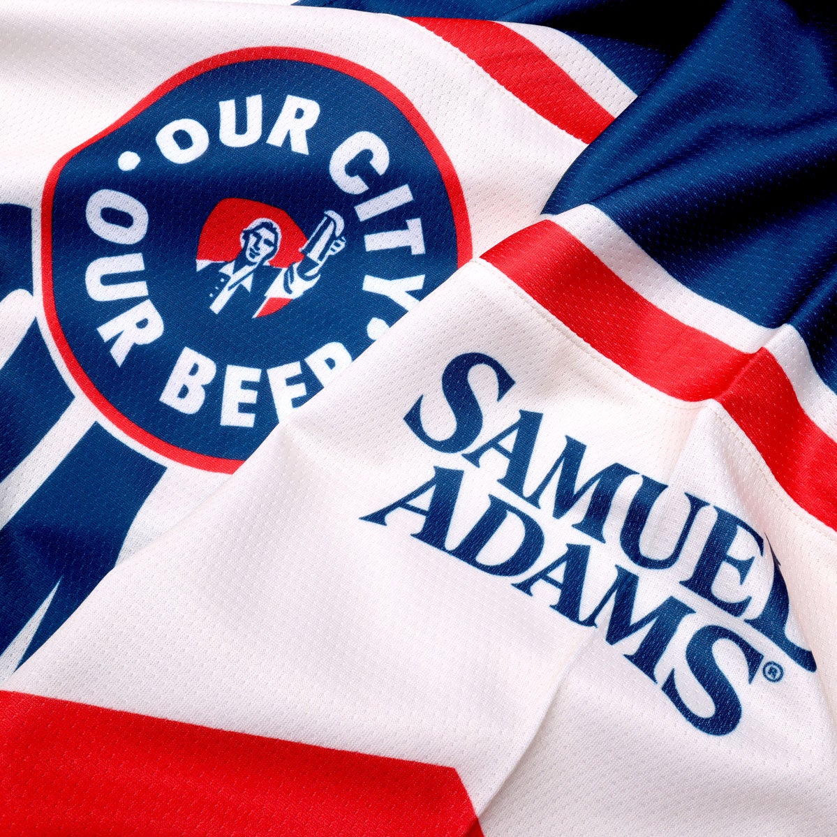 Base of jersey is white with blue and red patterns on the arms, waist, and shoulders. Centered "Boston" is written in blue, with a circle badge which features "Our City, Our Beer" and a red shield with Sam Adams hoisting a beer in the center of it. The Samuel Adams logo is featured in blue on the upper left arm.