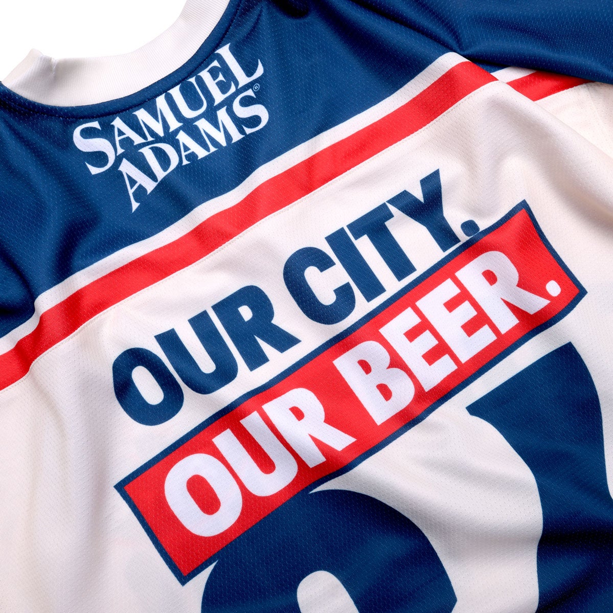 Base of jersey is white with blue and red patterns on the arms, waist, and shoulders. On the blue shoulder panel "Samuel Adams" is written in white. The 'name' on the jersey is "Our City Our Beer" which is written in half blue, half white. And the number on the jersey is 84. The Samuel Adams logo is featured on the left arm in blue.