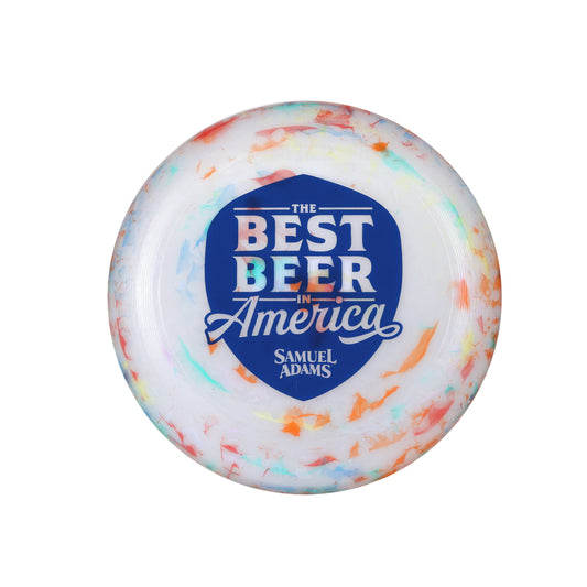 A frisbee with a white base but some multicolor accents, featuring a navy shield on the front with the phrase "The best beer in America" in it. Also features the Sam Adams logo.