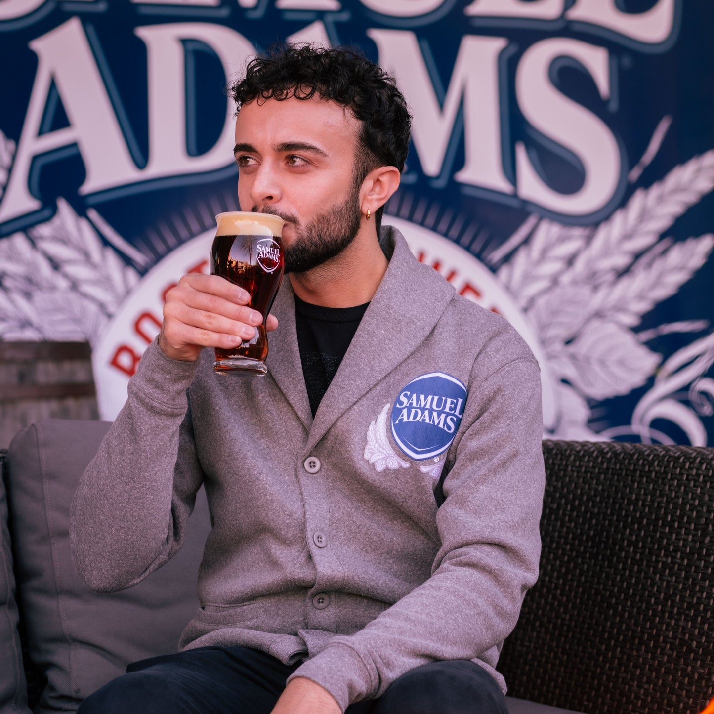 A Grey Henshaw Cardigan with button closures on the front and the classic Samuel Adams shield logo on the top left in navy and white. Pictured here is a man wearing the cardigan over a black tee shirt while sipping a perfect pint.