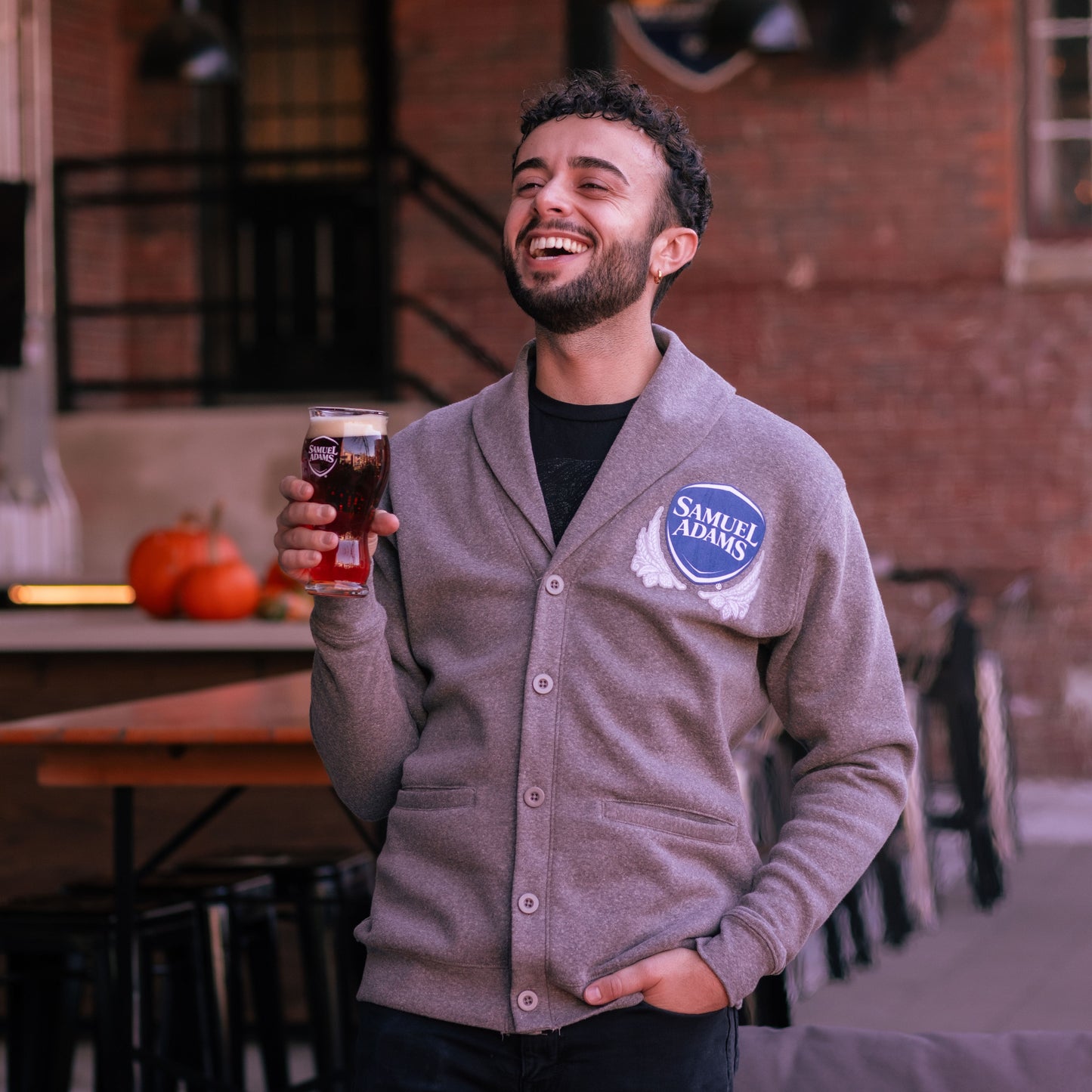A Grey Henshaw Cardigan with button closures on the front and the classic Samuel Adams shield logo on the top left in navy and white. Pictured here is a man wearing the cardigan over a black tee shirt while sipping a perfect pint.