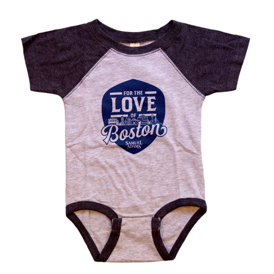 Grey onesie with navy shoulders and lining at the legs. A blue shield is featured on the center of the onesie with writing "For the Love of Boston" and the Samuel Adams logo under. The Boston skyline is featured within the writing.