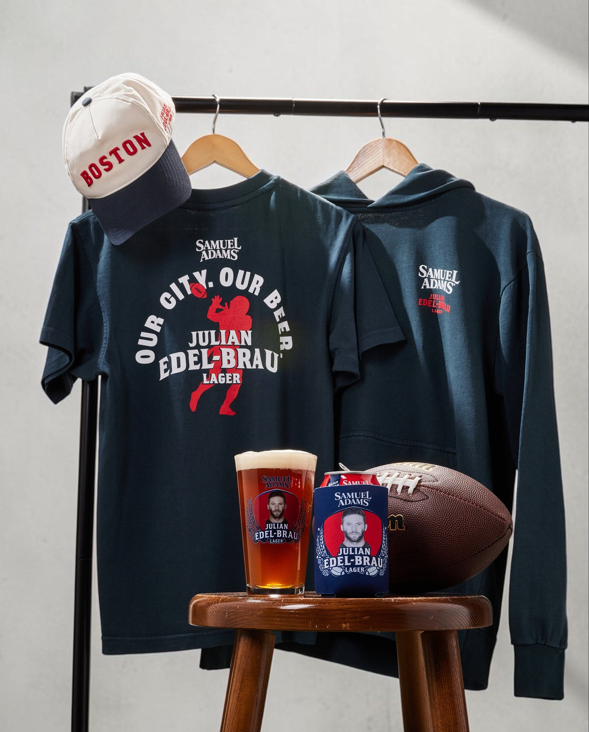 A styled apparel display featuring a navy blue t-shirt and matching hoodie hanging on a rack, both with red and white 'our City. our Beer' graphic prints. A white and navy Boston cap hangs to the left, and a wooden stool below holds a beer glass, a beer can, and a football.