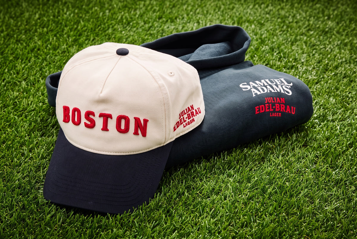 A navy blue baseball cap with 'BOSTON' in red on the front, resting on top of a folded navy hoodie on green grass. The hoodie features white and red embroidered text reading 'SAMUEL ADAMS JULIAN EDELBRAU'.
