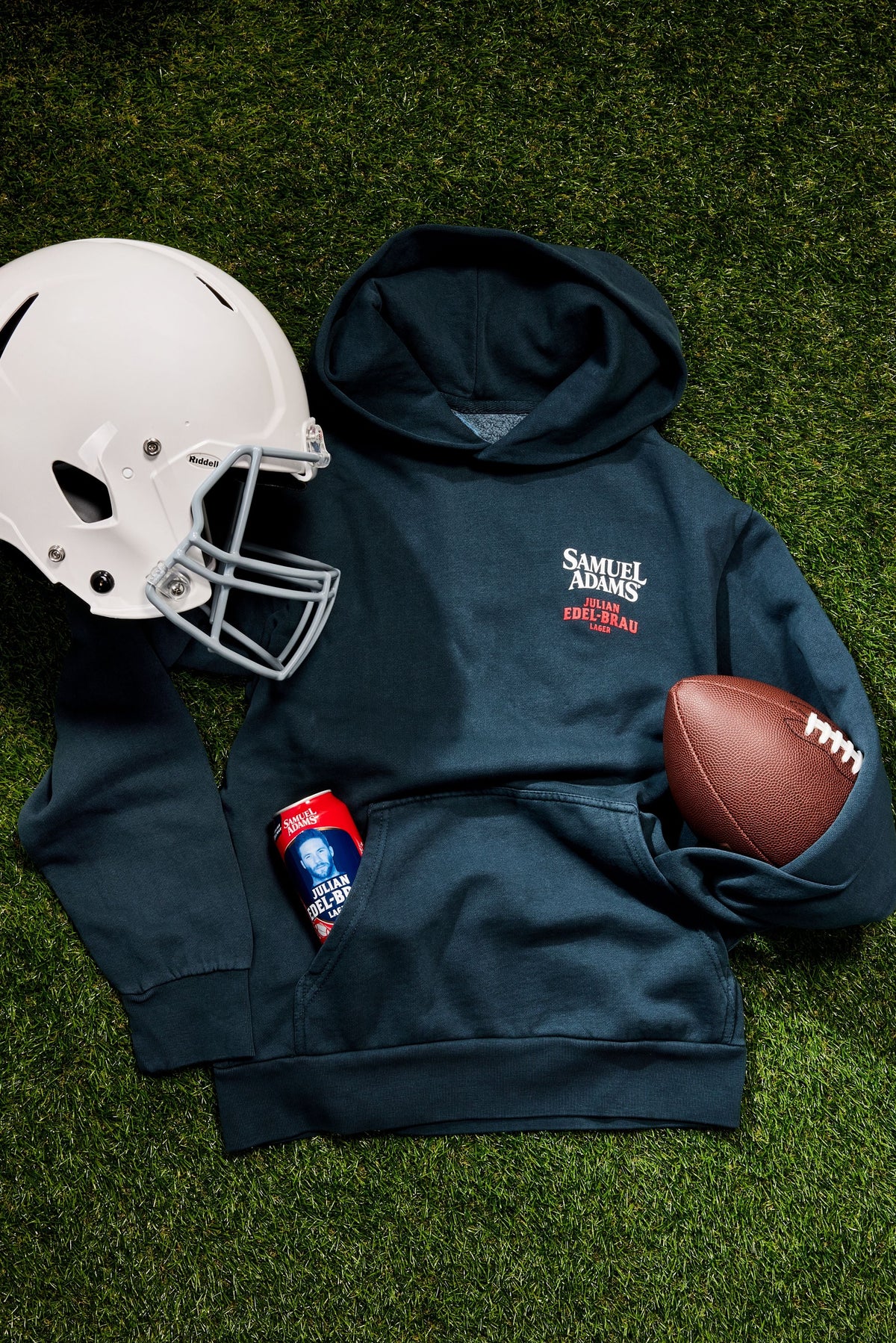 A navy blue hoodie laid flat on green turf with a white football helmet, a brown football, and a red beverage can styled around it. The hoodie has a white and red 'Samuel Adams' logo on the left chest.