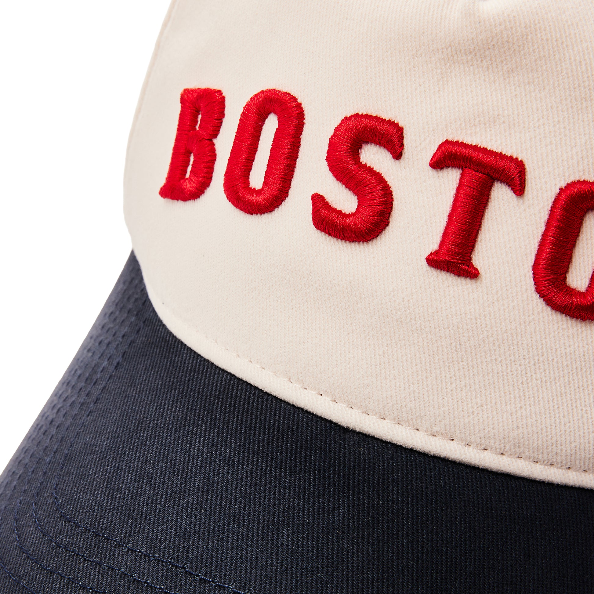 Close-up of a baseball cap with an off-white crown featuring red embroidered 'BOST' lettering and a dark navy blue brim.