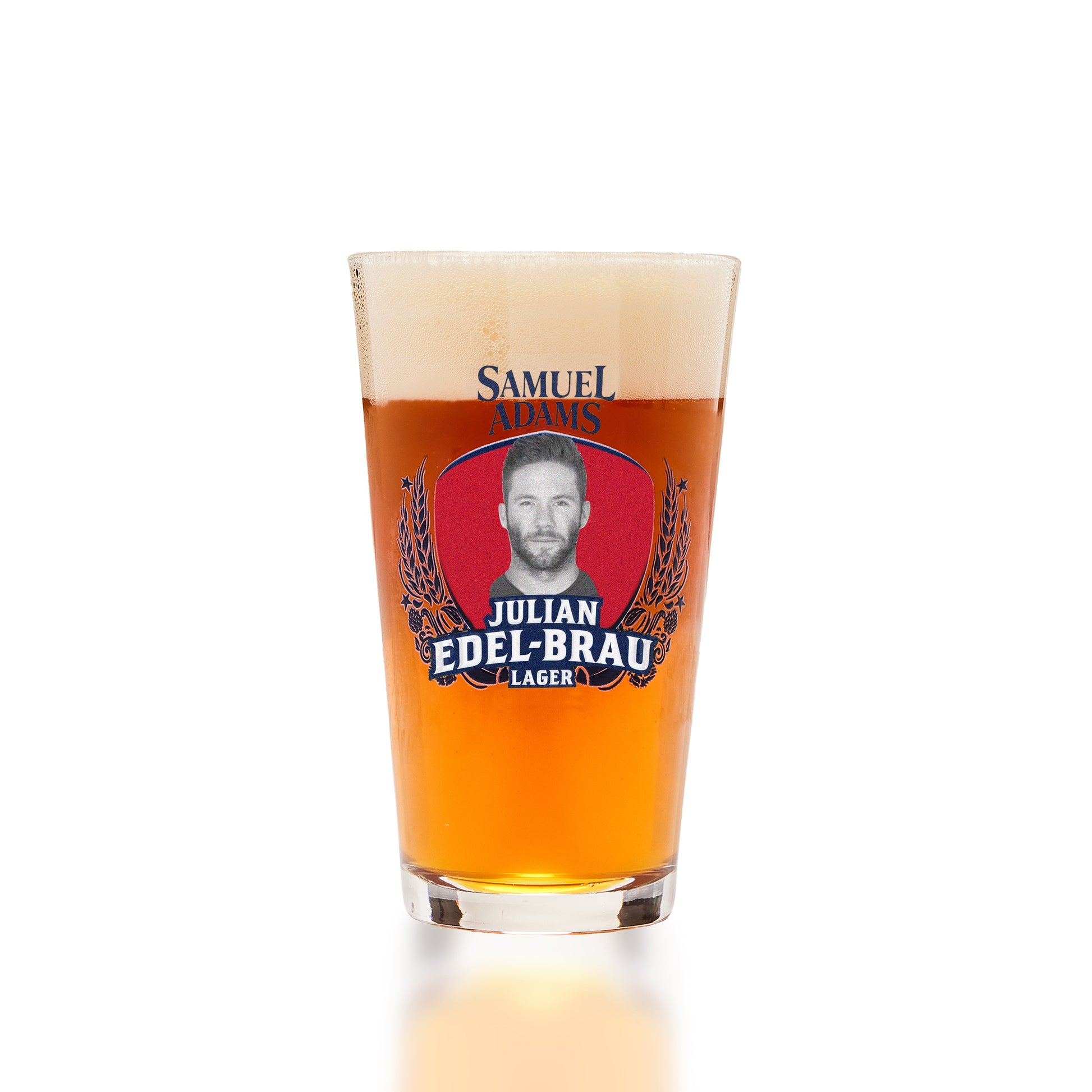 A clear pint glass filled with amber beer and topped with white foam, featuring a prominent Samuel Adams 'Julian Edel-Brau Lager' logo on the front, set against a clean white background.