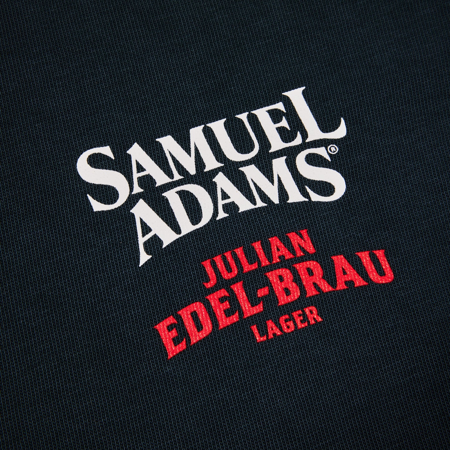 Close-up of a dark navy or black textured fabric with a large white 'SAMUEL ADAMS' logo and red 'JULIAN EDEL-BRAU LAGER' text printed on it.