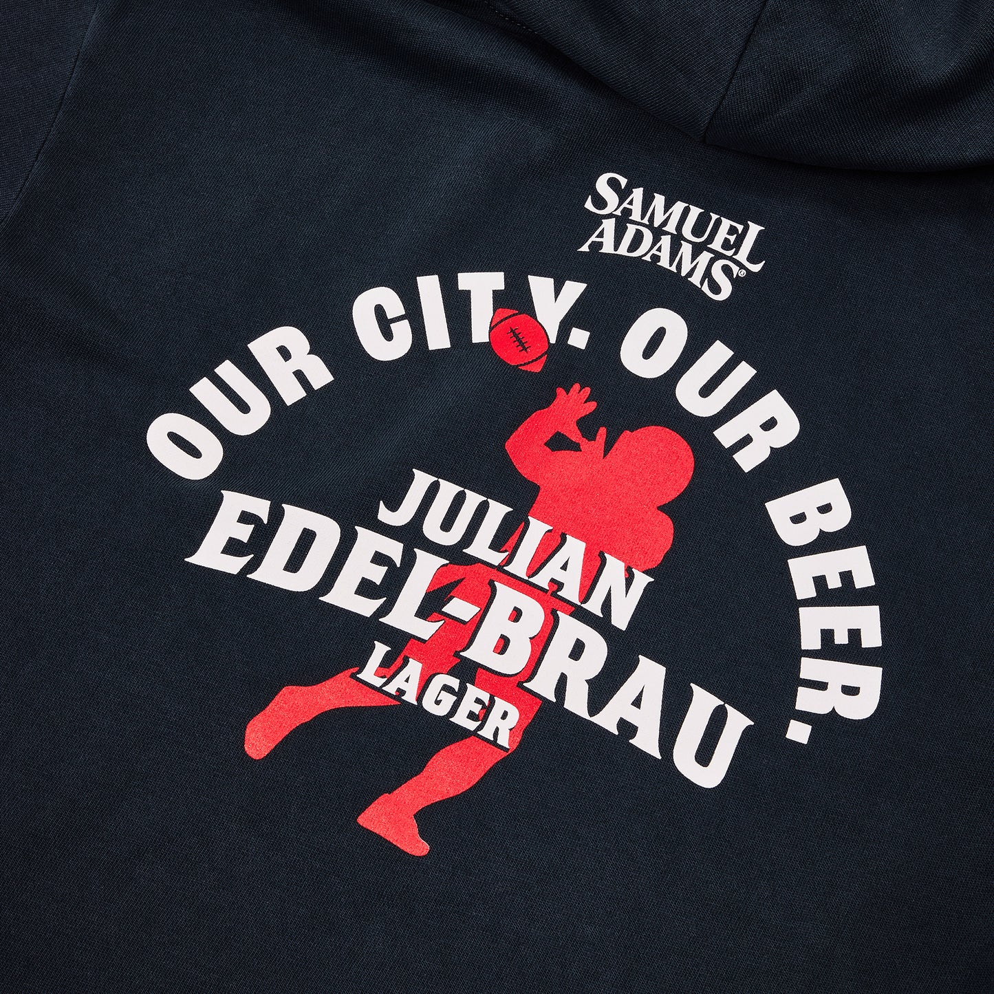 Close-up of a dark navy hoodie featuring a custom graphic print. The design includes a red silhouette of a football player, a football, and bold white text reading 'OUR CITY. OUR BEER.'
