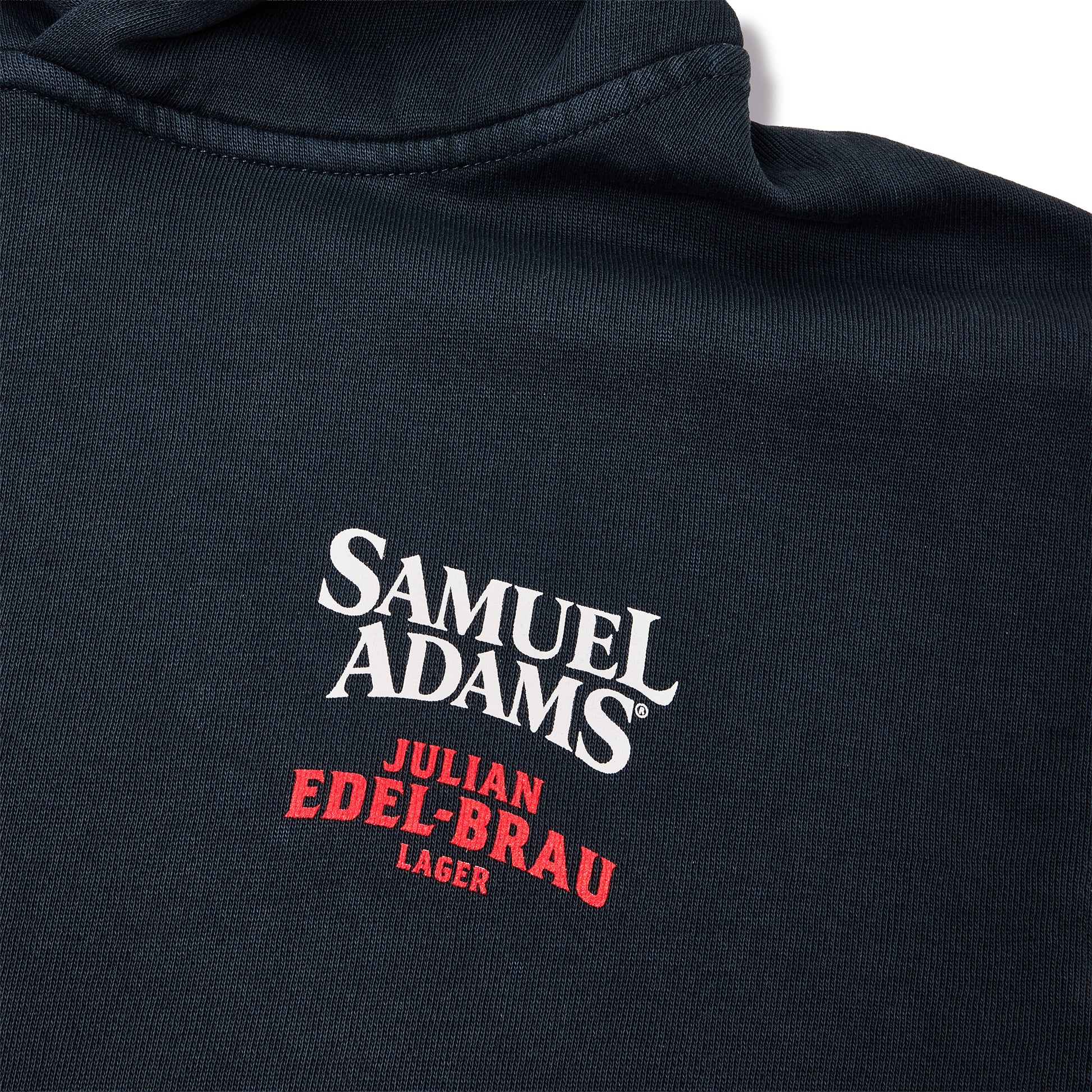 Close-up of a dark navy blue fabric surface with embroidered text reading 'SAMUEL ADAMS' in white and 'JULIAN EDEL-BRAU LAGER' in red.