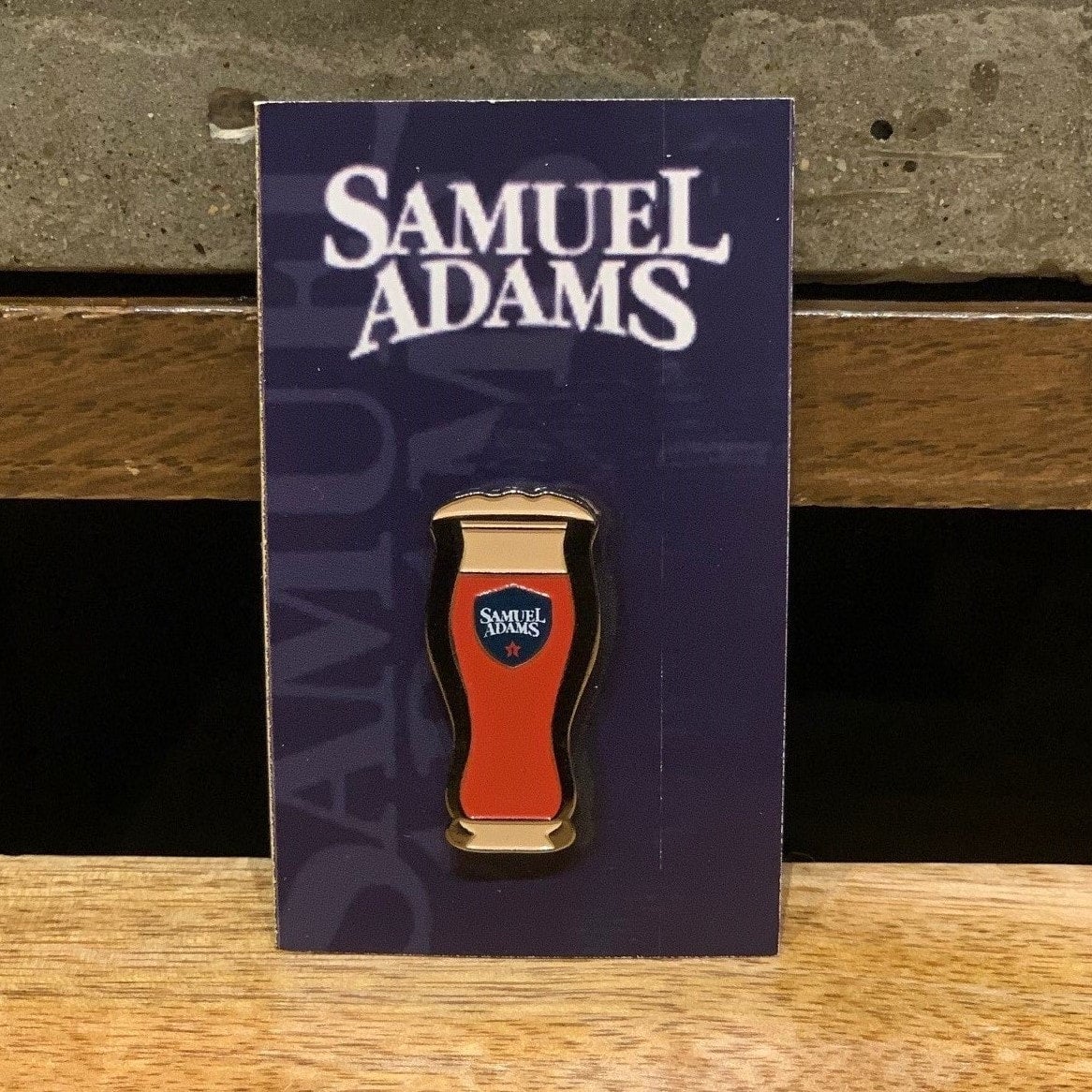 A cast alloy metal pin modeled after a perfect pint of Sam Adams beer with metal clasp on the back. Pictured here on a carboard backer.