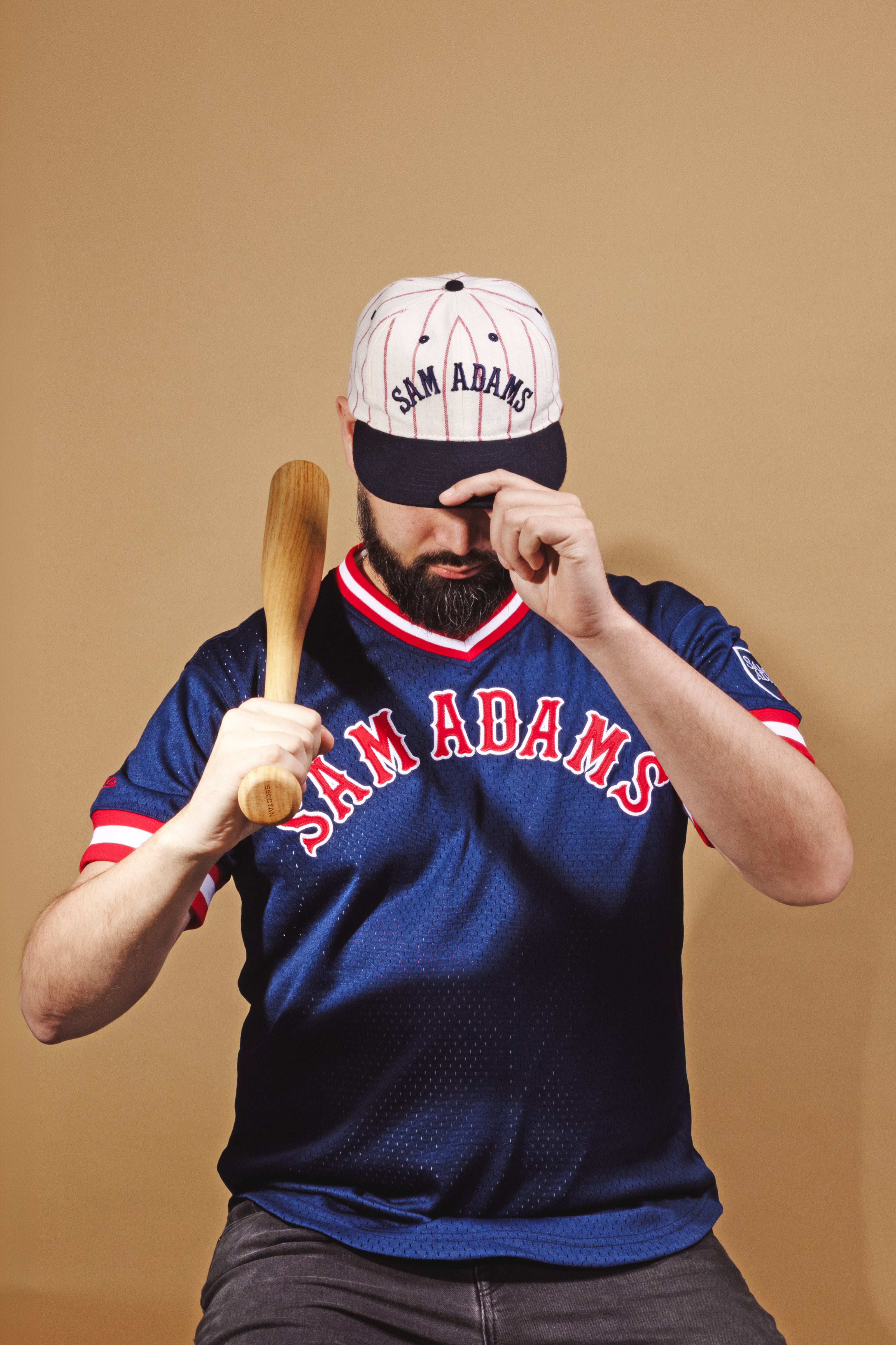 Person wearing a Sam Adams baseball cap and jersey, holding a bat against a beige background