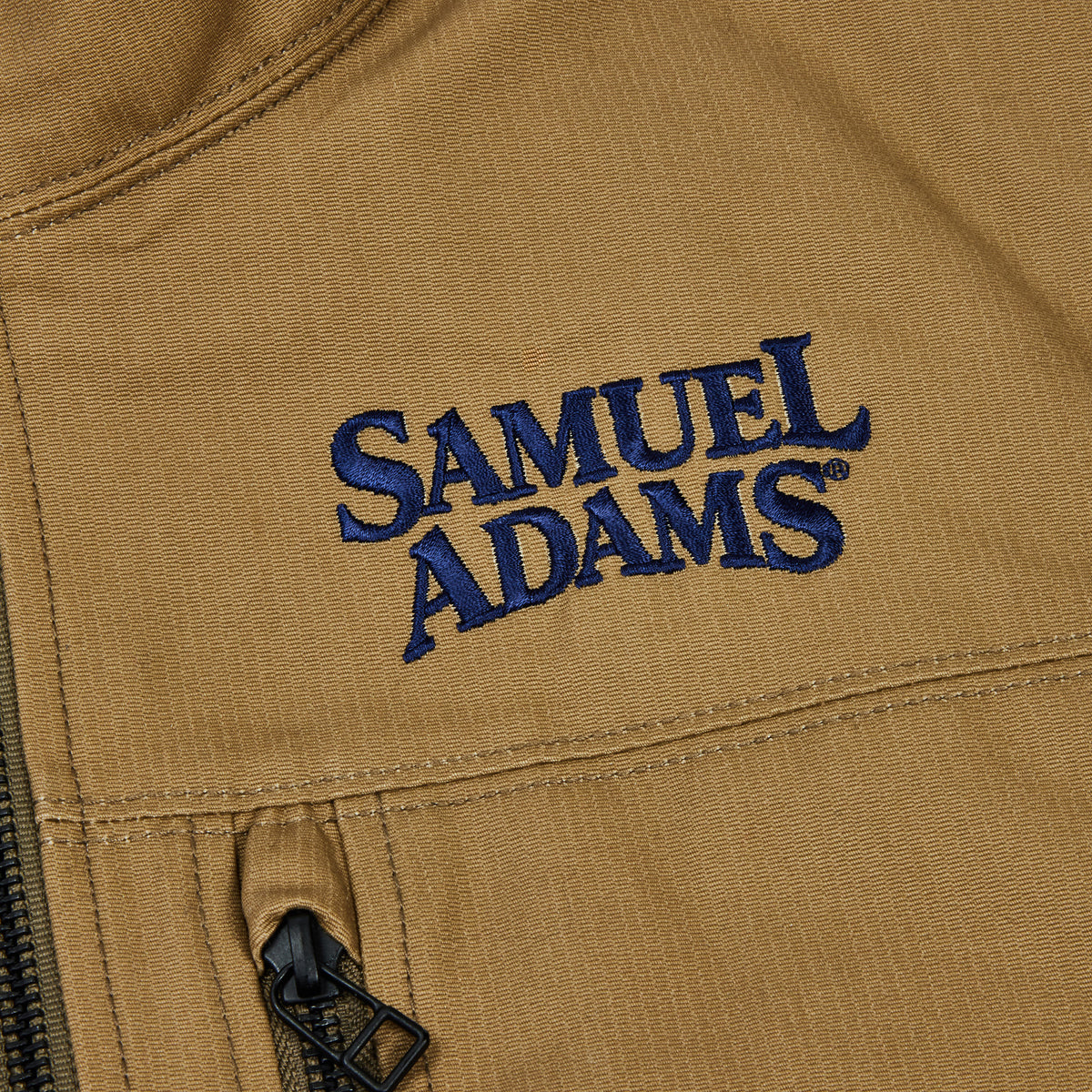 Close-up of a tan textured fabric jacket with a dark blue embroidered 'SAMUEL ADAMS' logo and a black zipper pull.