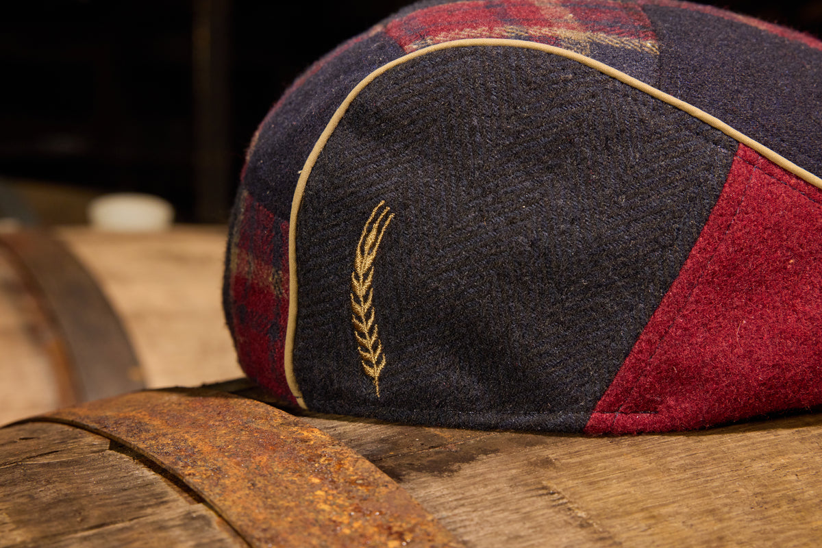 A close-up of a Scally flat cap with a dark navy blue body, red and black plaid side panels, a gold embroidered wheat stalk, and gold piping, resting on a rustic wooden barrel.