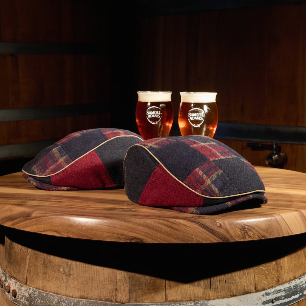 Two tartan plaid scally caps rest on the top of a wooden barrel, with two glasses of amber beer behind them against a dark wood barrel room background.