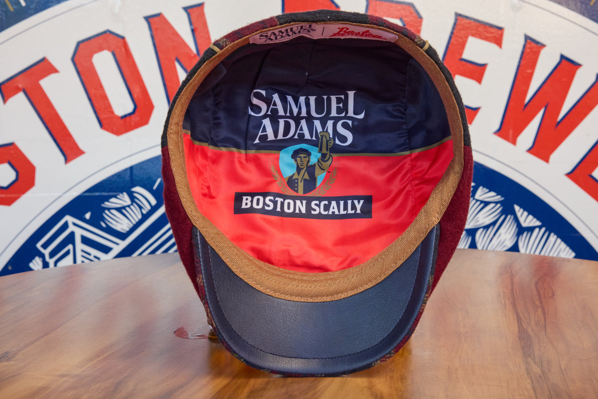 A vintage Boston Scally cap with a dark red and navy blue front panel, a tan visor trim, and a navy blue brim, displayed on a wooden table. The inside of the crown features a red and navy blue lining with 'SAMUEL ADAMS' and 'BOSTON SCALY' embroidered text.