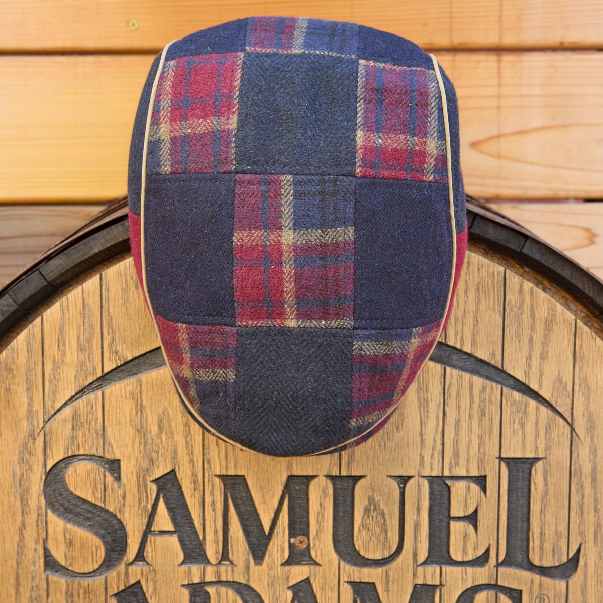 A patchwork tweed flat cap with a navy blue, red, and beige plaid pattern, displayed on top of a wooden barrel with the Samuel Adams logo.