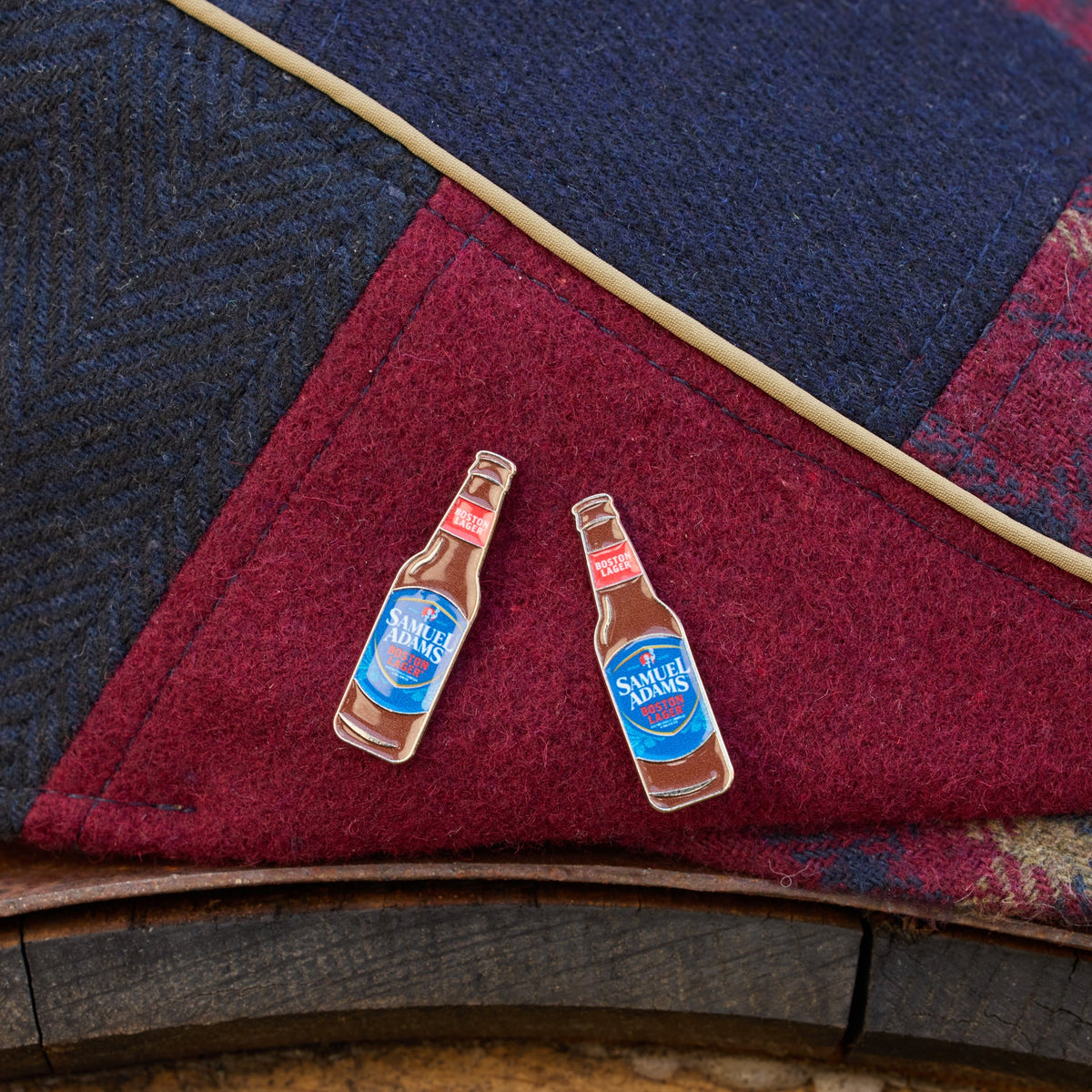 Two enamel lapel pins shaped like miniature bottles of Samuel Adams beer, complete with blue labels and red caps, displayed on a dark red and navy blue herringbone wool fabric background.
