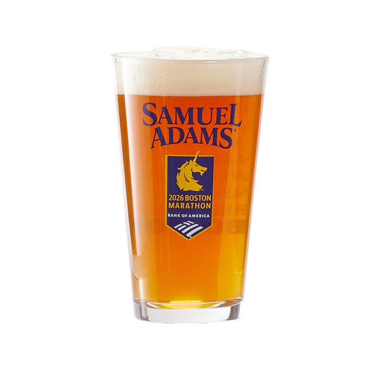 A pint glass filled with amber-colored beer topped with a foamy head. The glass features the Samuel Adams logo in orange and a blue emblem below it that reads “2026 Boston Marathon – Bank of America,” along with a stylized unicorn graphic
