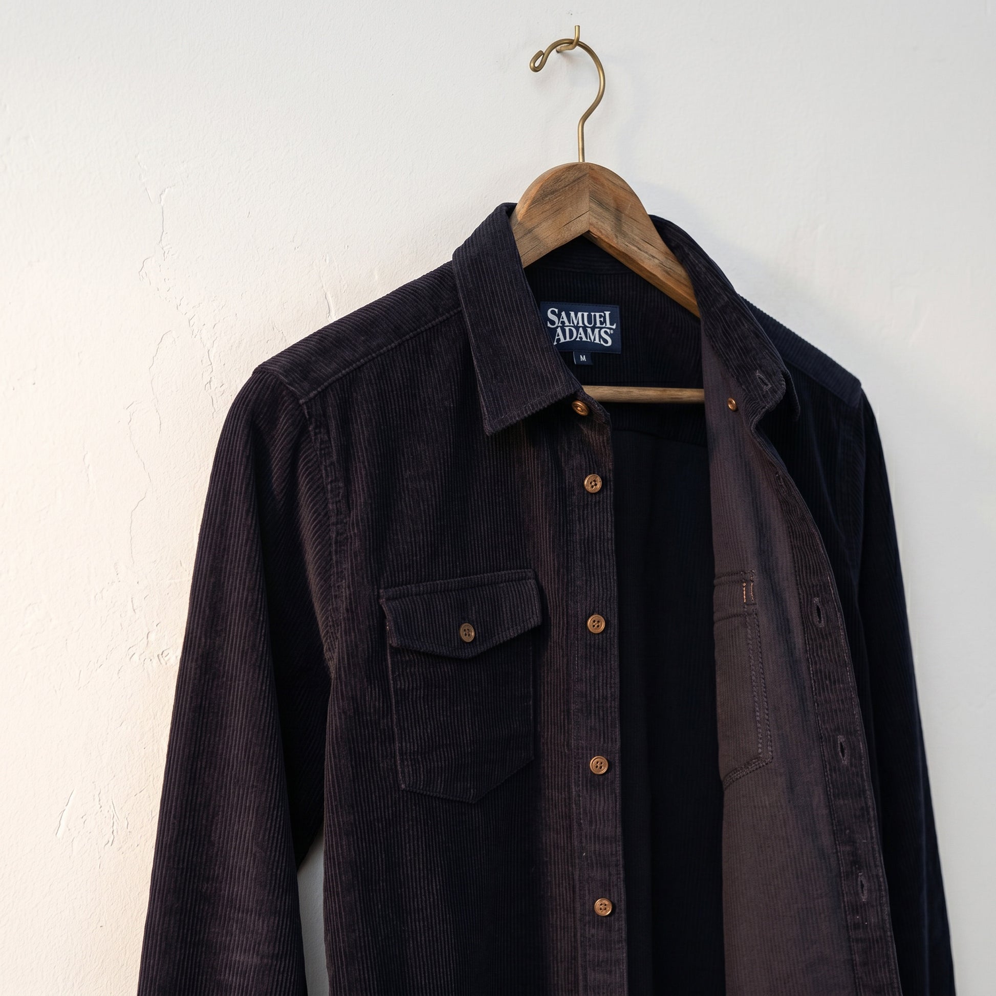 An angled view of a navy button down corduroy flannel shirt,, unbuttoned on a hanger against a white background. The Sam Adams logo is written in white on the tag. 
