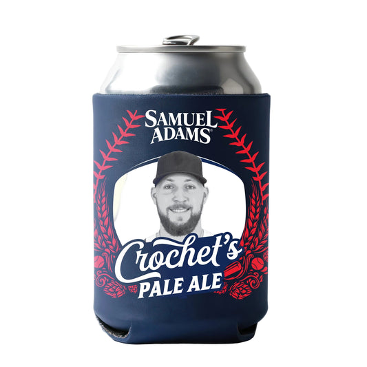 A silver can wrapped in a navy can cooler featuring red baseball stitching graphics, the Samuel Adams logo, and the text “Crochet’s Pale Ale” in bold lettering, with a central oval frame black and white headshot of Garrett Crochet  and decorative red illustrations.