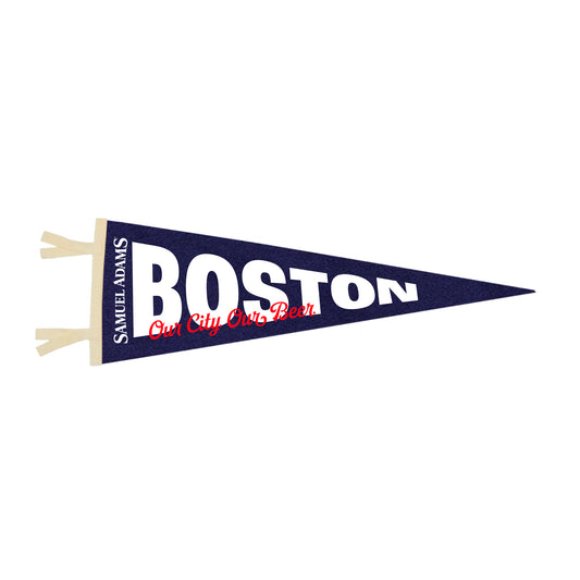 A navy blue felt pennant with a tapered triangular shape featuring bold white text reading “BOSTON” across the center. Below the city name, stylized red script displays the phrase “Our City Our Beer.” The left edge includes a vertical white strip with “Samuel Adams” printed in white, along with two cream‑colored tie tabs for hanging. 