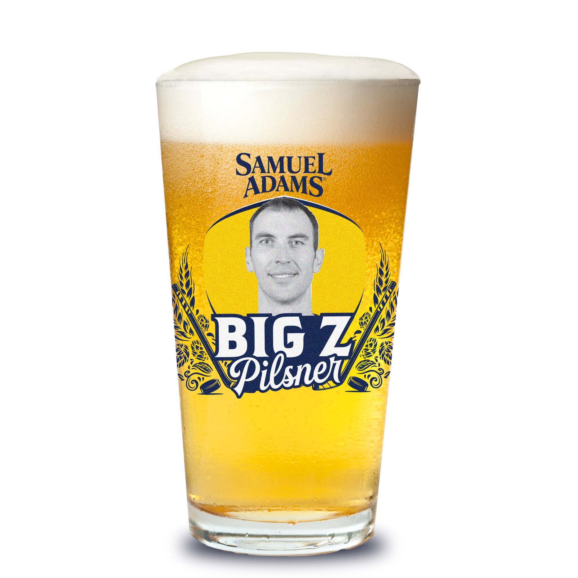 A Samuel Adams pint glass filled with golden beer and topped with foam, featuring a blue‑and‑yellow label framed by barley graphics; at center is a black‑and‑white portrait of Big Z above bold text that reads “Big Z Pilsner,” with the Samuel Adams wordmark above the label.