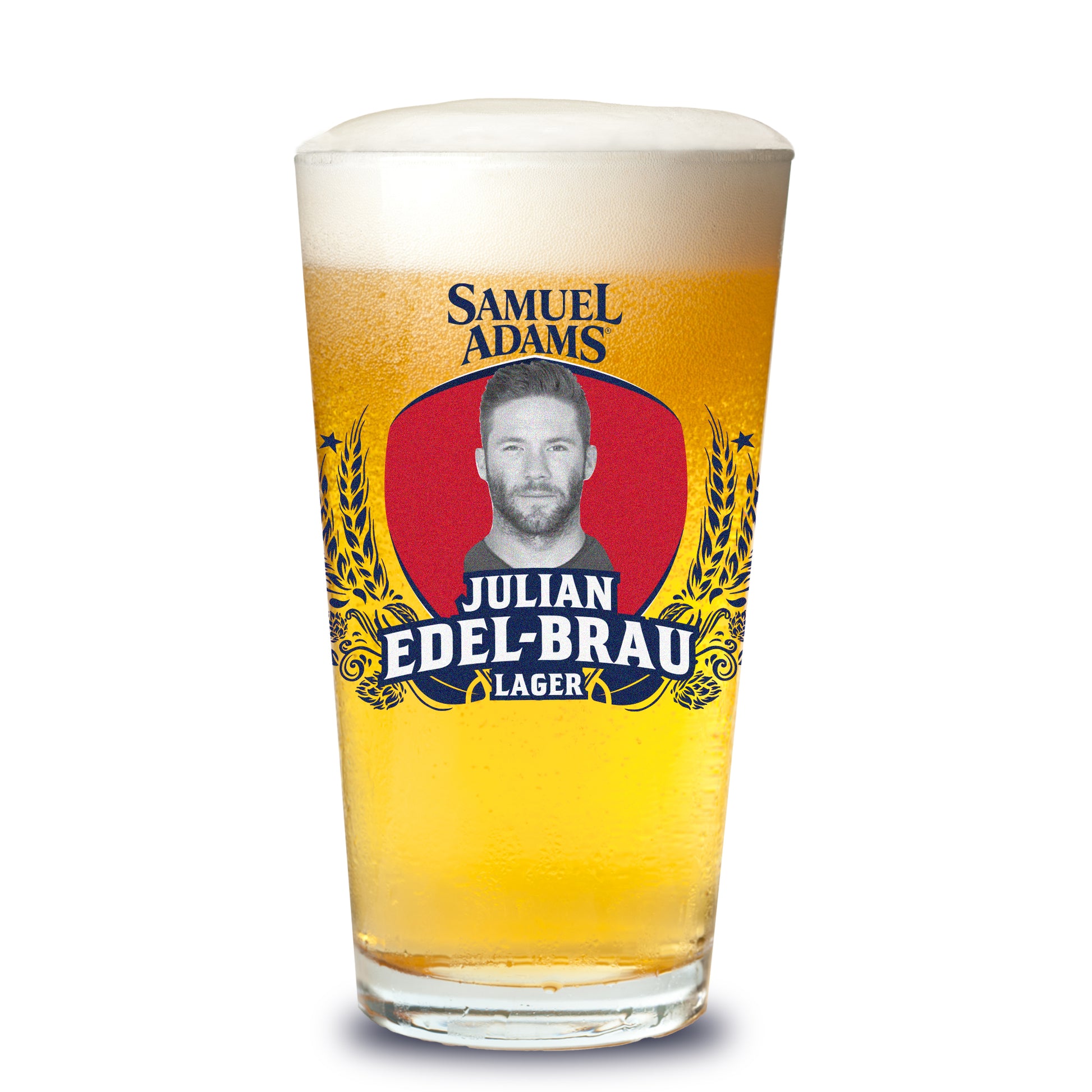 A Samuel Adams pint glass filled with golden beer and topped with foam, featuring a red oval label flanked by golden barley illustrations; at center is a black‑and‑white portrait of Julian Edelman, with stylized text beneath that reads “Julian Edel‑Brau Lager,” and the Samuel Adams wordmark above.