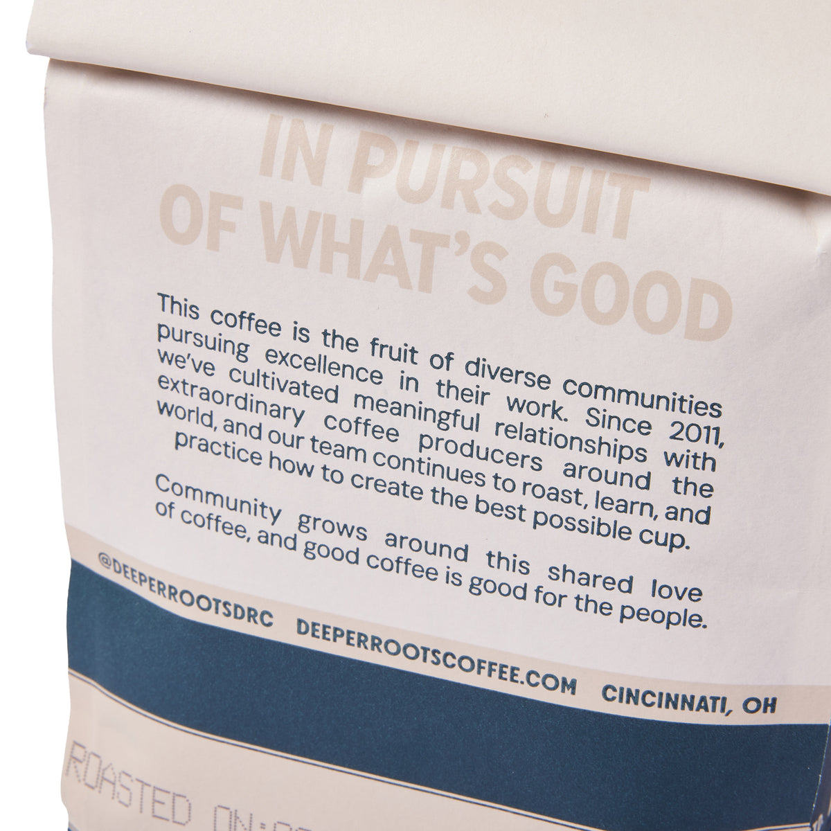 Close-up of a coffee bag or package with printed text. The visible design has a cream background, large muted tan headline text reading “IN PURSUIT OF WHAT’S GOOD,” a block of smaller black descriptive text, and a dark navy band near the bottom with the brand name DEEPERROOTS COFFEE and “CINCINNATI, OH.