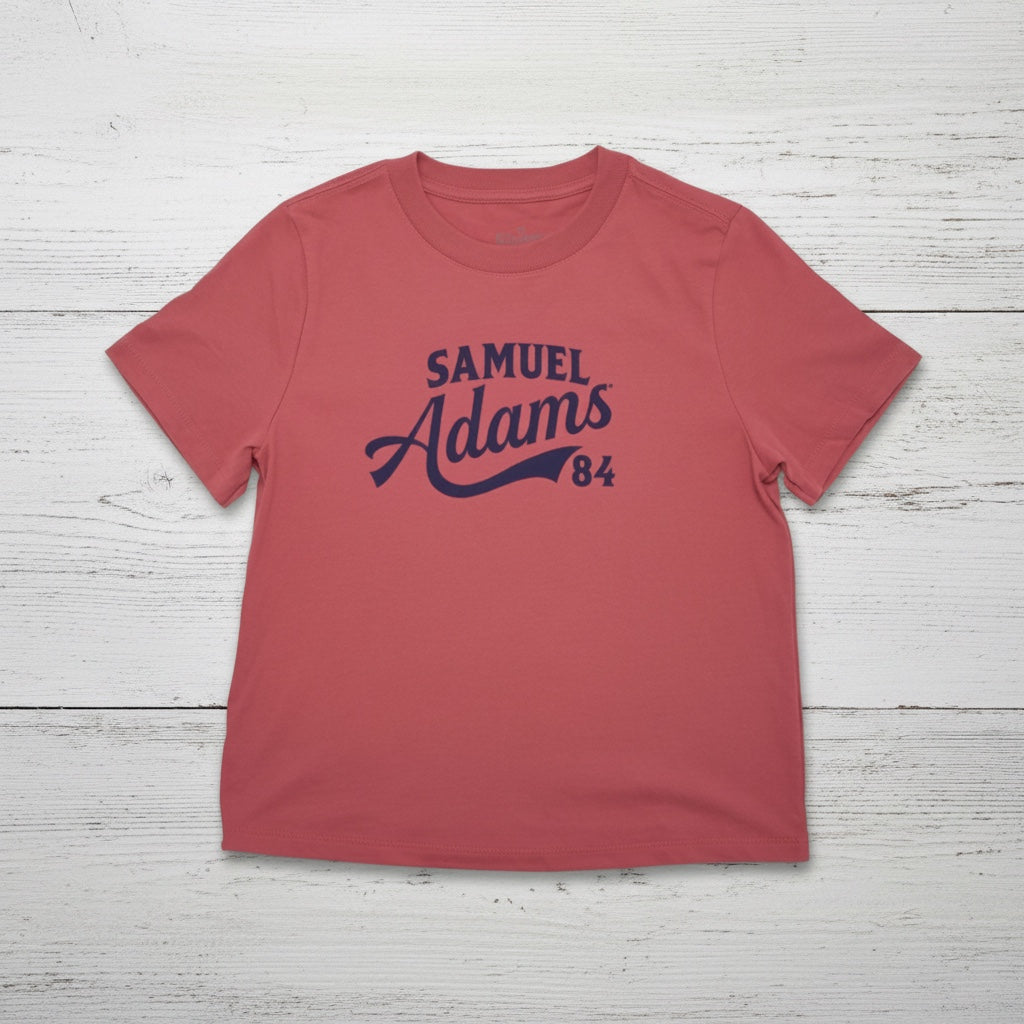 A pale red tee shirt against a white wooden background. "Samuel Adams 84" is written in a blue sport logo font across the front. 