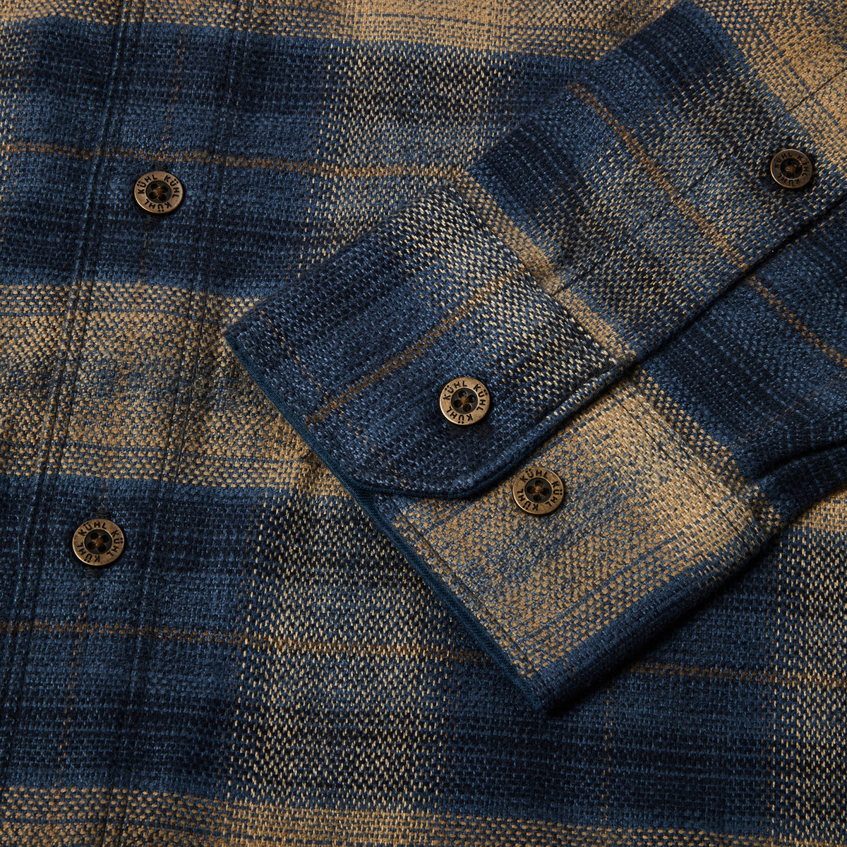 Close-up of a blue and tan plaid button-up shirt fabric with a folded cuff showing dark brown buttons.