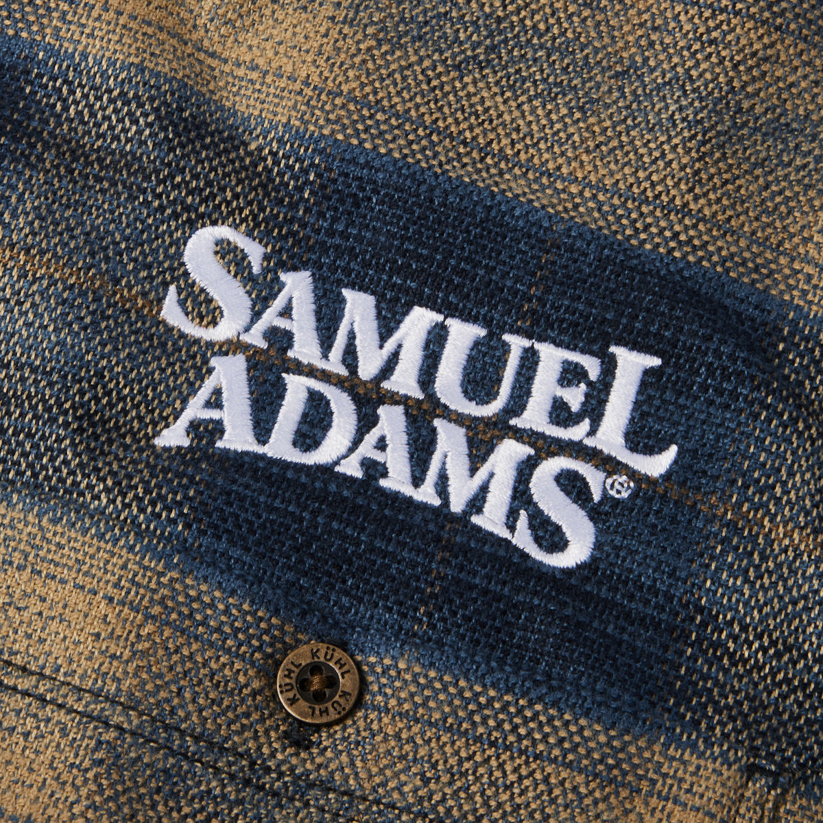 Close-up of a textured flannel shirt fabric with a blue and tan plaid pattern, featuring a white embroidered 'SAMUEL ADAMS' logo and a brown button.