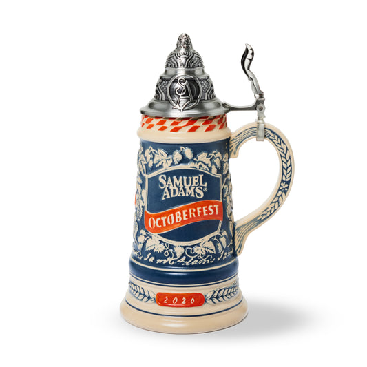 A ceramic Oktoberfest stein with a pewter lid and handle, featuring blue and white floral patterns, a red banner reading 'Samuel Adams Oktoberfest', and '2026' on the base, shown on a white background.
