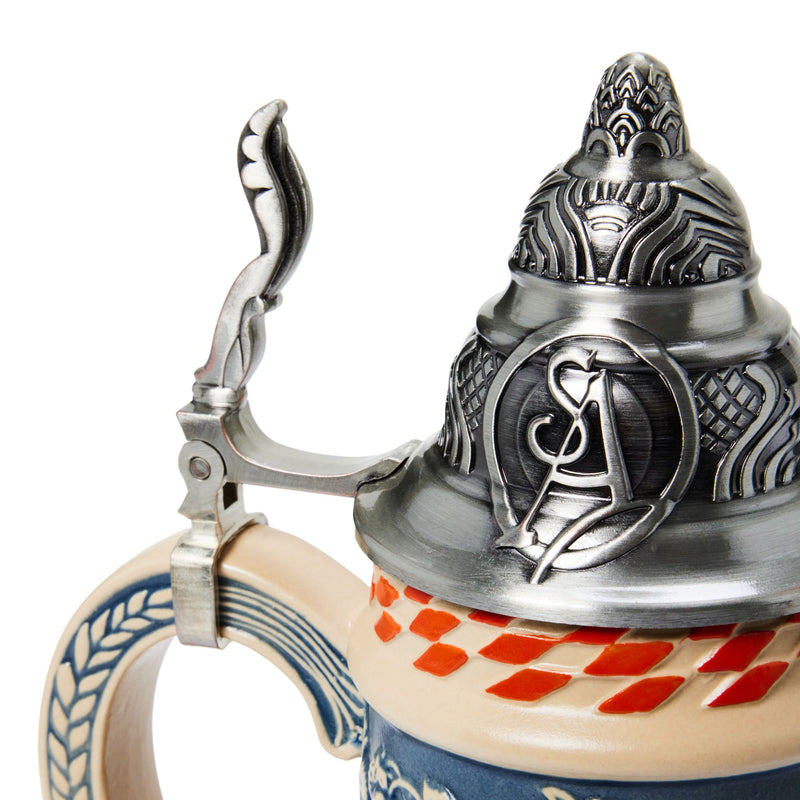 Close-up of a decorative beer stein with a silver pewter lid featuring embossed relief details and a pewter handle, attached to a beige ceramic body with blue and red diamond patterns.