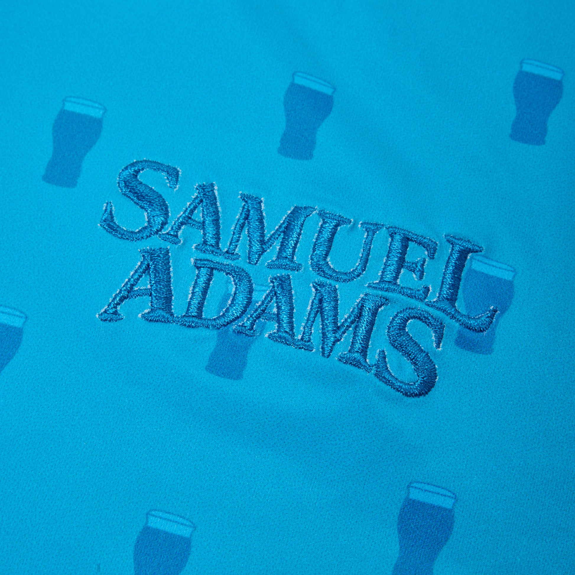 A close-up of a bright blue fabric surface featuring the embroidered text 'SAMUEL ADAMS' in dark blue thread, surrounded by small embossed beer glass motifs.