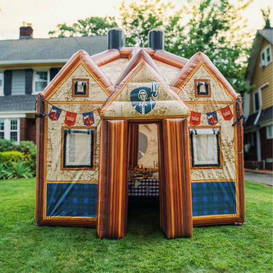 An inflatable beer hall-themed house blowup set up on a green lawn in front of houses, featuring a beige and brown wood-grain exterior with blue plaid accents and a Samuel Adams image over a opening to enter the inflatable