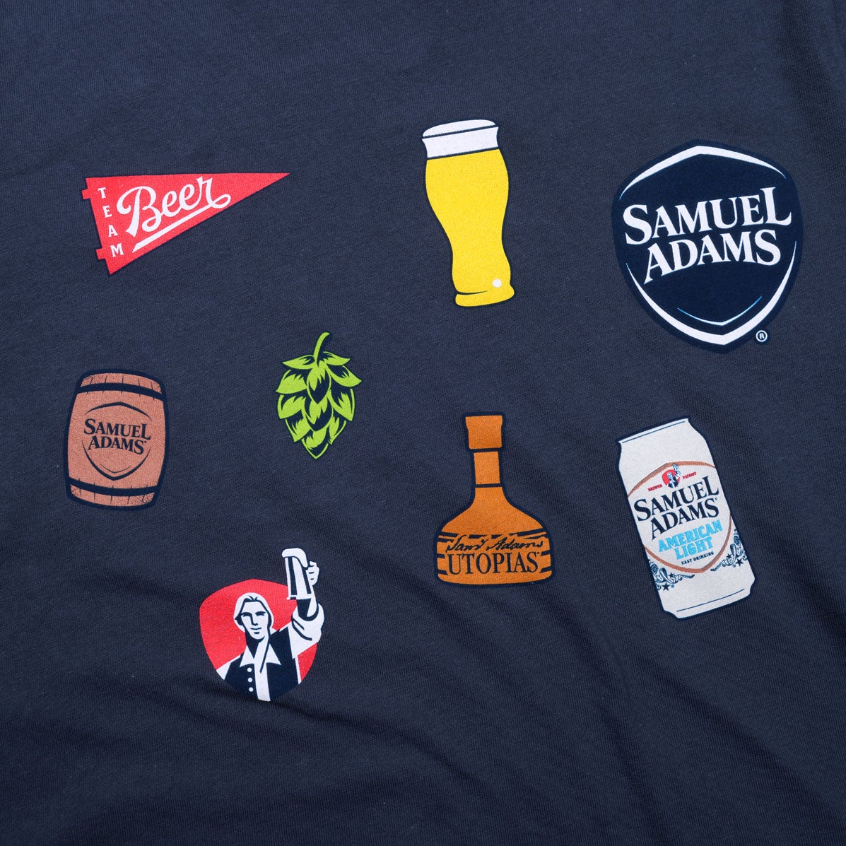 Close up of the clip art on the front of the tee. Featuring a red beer banner, a pint of beer, the Sam Adams crest logo, a Sam Adams barrel, a hop plant, the Utopias bottle, a Sam Adams can, and a red crest with Sam Adams hoisting a beer.