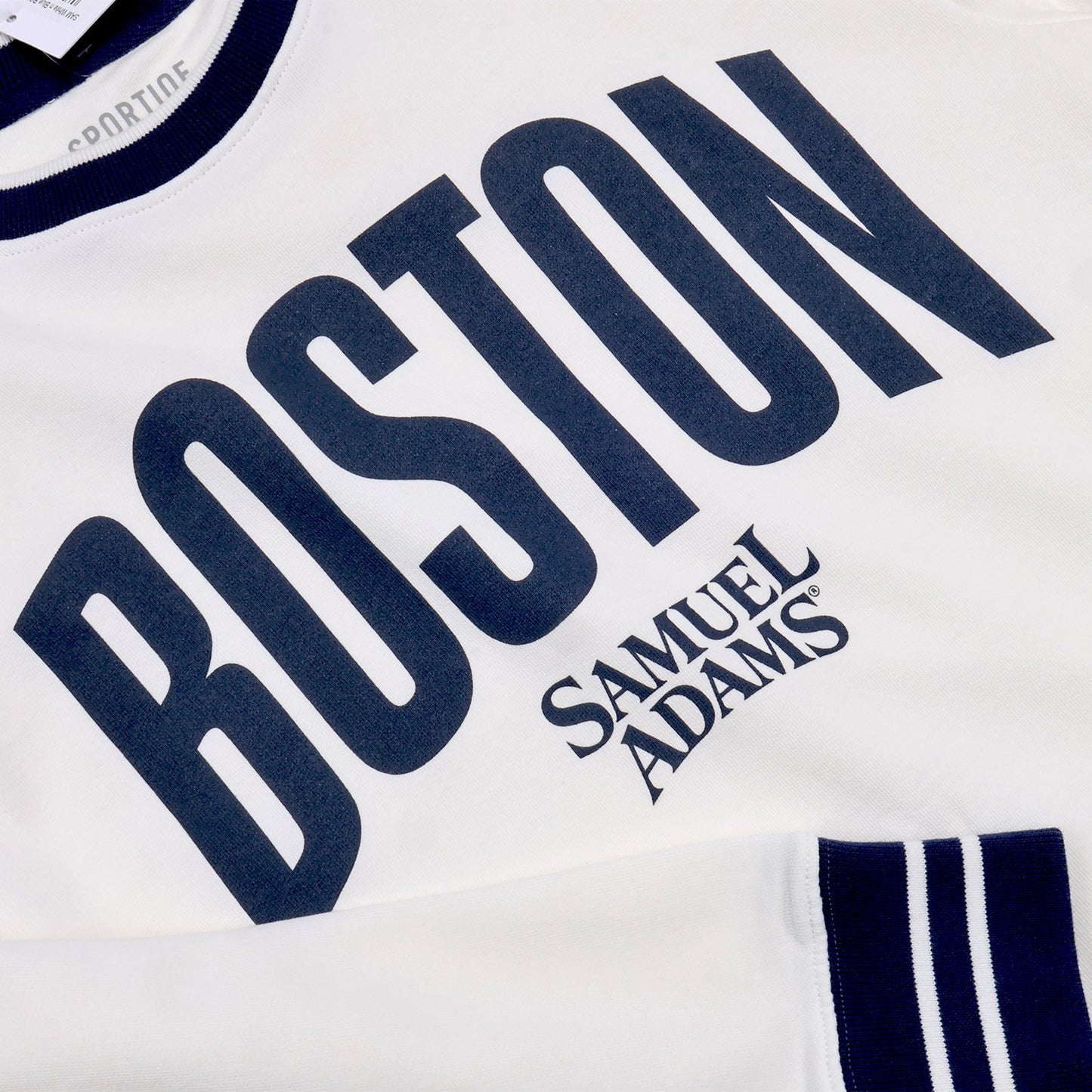 Close up of a white crewneck sweater with thick navy stripes around the neckband, cuff, and hem of the sweater. In matching large navy text reads "BOSTON", beneath in smaller text in the Samuel Adam's logo. Close up shows the "Sportiqe" brand logo printed on the tag of the sweater in grey.