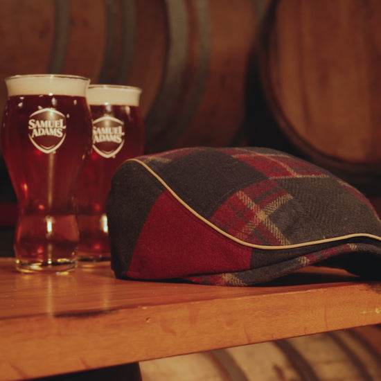 Video showing plaid and navy Scally cap on a table with two pints of beer to the left.  The shelf is floating amongst oak barrels in the background.