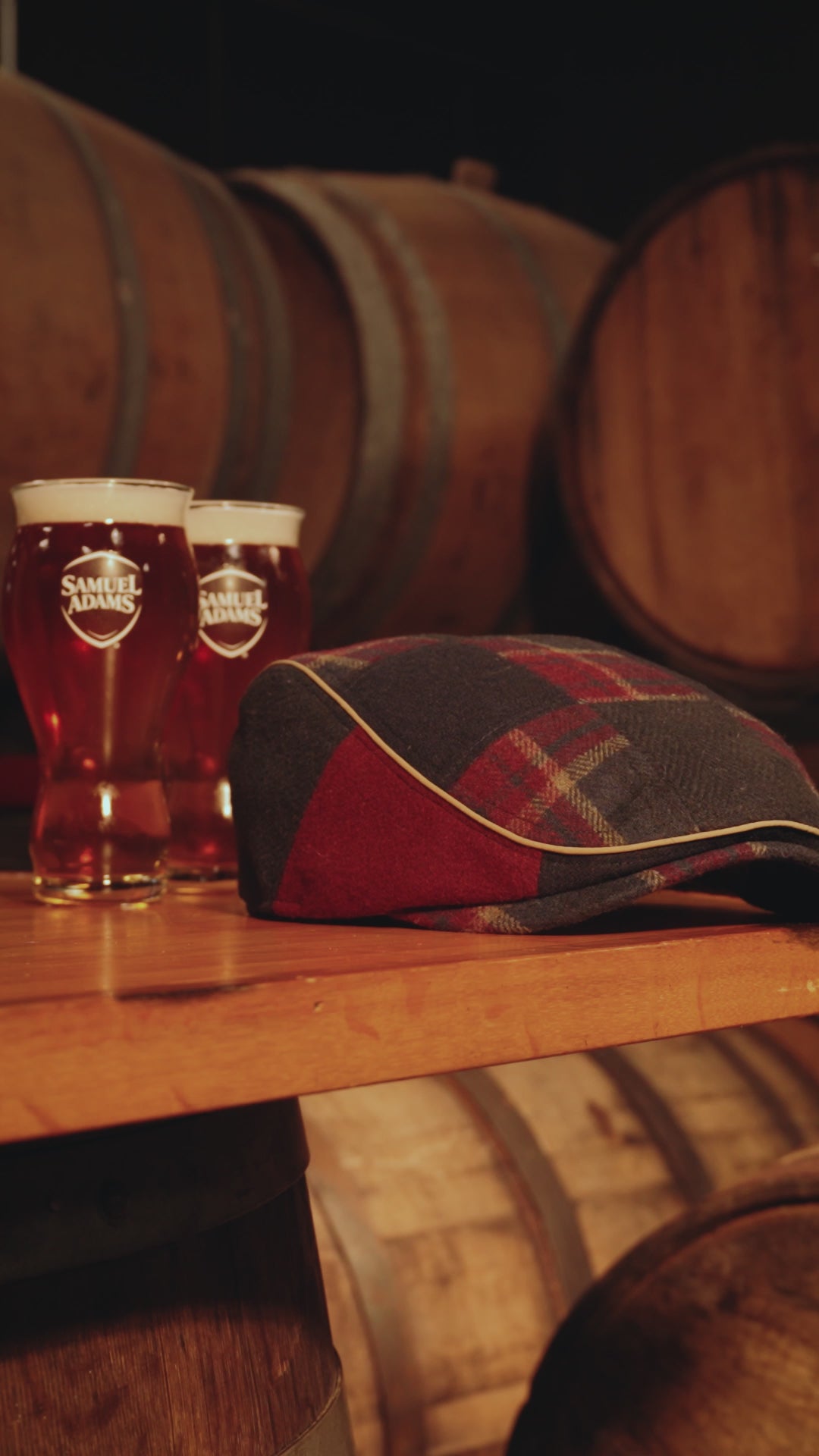 Video showing plaid and navy Scally cap on a table with two pints of beer to the left.  The shelf is floating amongst oak barrels in the background.